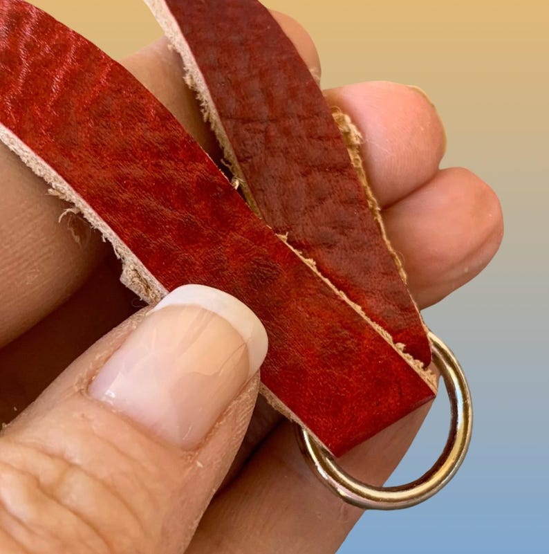 Op de afbeelding: Close-up van een rode leren riem met een goudkleurige metalen ring. Het leer heeft een textuuroppervlak en wordt op zijn plaats gehouden door een metalen ring.