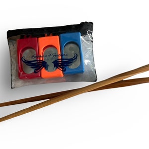 Op de afbeelding: Een doorzichtig plastic zakje met drie gekleurde eetstokjeshulpen in rood, oranje en blauw. Twee houten eetstokjes zijn eronder geplaatst. De tekst "Leachers 4 Feathers" is zichtbaar op het zakje.
