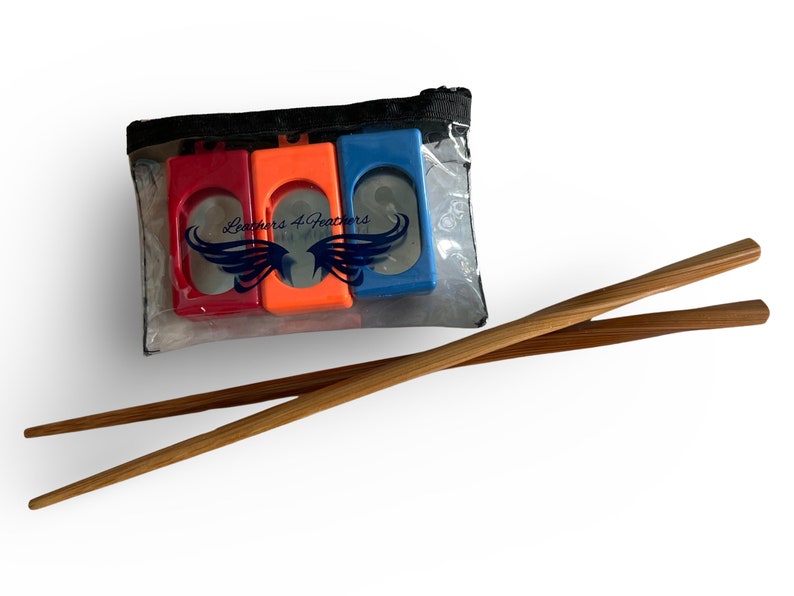 Op de afbeelding: Een doorzichtig plastic zakje met drie gekleurde eetstokjeshulpen in rood, oranje en blauw. Twee houten eetstokjes zijn eronder geplaatst. De tekst "Leachers 4 Feathers" is zichtbaar op het zakje.