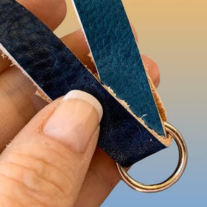 Op de afbeelding: Close-up van een leren sleutelhanger met een goudkleurige ring. De sleutelhanger heeft twee stroken blauw leer, &eacute;&eacute;n donker marineblauw en de andere teal, verbonden door de ring. De randen van het leer zijn rauw en onafgewerkt.