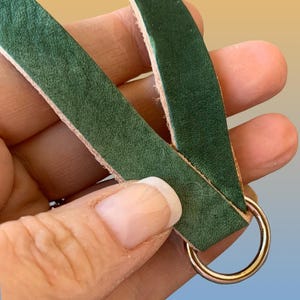 Op de afbeelding: Close-up van een groene leren riem met een goudkleurige metalen ring. Het leer heeft een getextureerde uitstraling en wordt in een hand gehouden. De ring is cirkelvormig en glanzend.