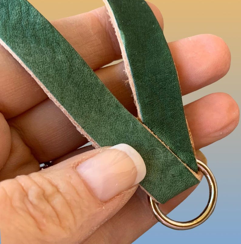 Op de afbeelding: Close-up van een groene leren riem met een goudkleurige metalen ring. Het leer heeft een getextureerde uitstraling en wordt in een hand gehouden. De ring is cirkelvormig en glanzend.