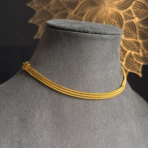 May include: A gold necklace with a woven design is displayed on a grey mannequin. The necklace has a flat, multi-strand design. The background is a dark colour with a gold floral pattern.