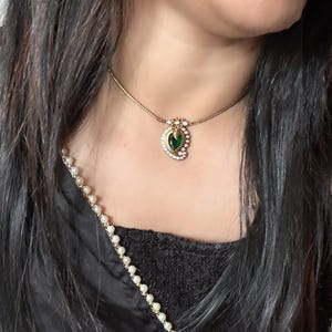 May include: A gold necklace with a pendant. The pendant has a green gemstone at its centre, encircled by small, clear stones. The necklace is worn against a black top. The chain is delicate.
