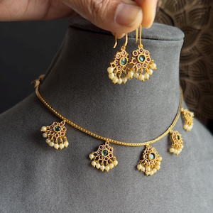May include: Gold-toned necklace and earrings set. The necklace features a delicate chain with multiple pendant charms. Each charm and the earrings have a floral design with green and red accents, and small white pearls. The jewelry is displayed on a gray mannequin.