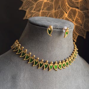 May include: Gold-coloured necklace and matching earrings set. The necklace features a series of green leaf-shaped pendants. The earrings mirror the leaf design. The jewellery is displayed on a grey bust against a dark background.