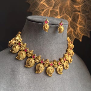 May include: A gold-coloured necklace and earring set. The necklace has ornate, paisley-shaped pendants with red gemstone accents. The earrings echo the design with similar gold detailing and red stones.