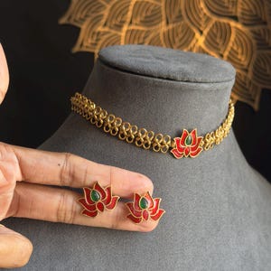 Puede incluir: Conjunto de collar y pendientes dorados. El collar y los pendientes presentan un diseño de flor de loto con pétalos de esmalte rojo y un centro verde. La joyería se exhibe sobre una cabeza de maniquí gris.