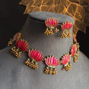 Puede incluir: Un conjunto de collar y pendientes dorados con colgantes de flores de loto rosas y rojas con detalles verdes. Las joyas se exhiben sobre un busto gris, con un patrón dorado decorativo en el fondo.