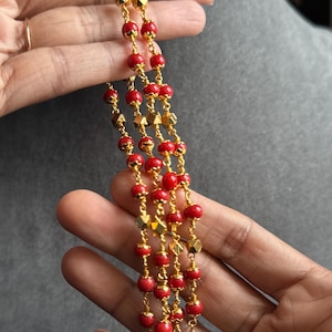 Puede incluir: Un collar dorado con tres hilos de cuentas rojas y detalles geométricos dorados. El collar se sostiene en una mano, mostrando el diseño y el color de la joya.
