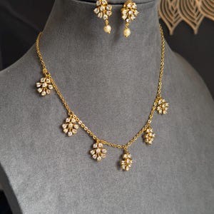 Puede incluir: Un conjunto de collar y pendientes en tono dorado con delicados dijes en forma de flor incrustados con cristales blancos transparentes. La cadena del collar tiene cinco colgantes florales espaciados, y cada pendiente presenta una flor con una pequeña perla blanca colgante.