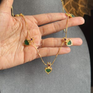 May include: Gold-coloured necklace with green and red gemstone accents. The necklace features a central pendant and two side pendants, all with a heart-shaped green stone and a small red stone. The necklace is held against a grey background.