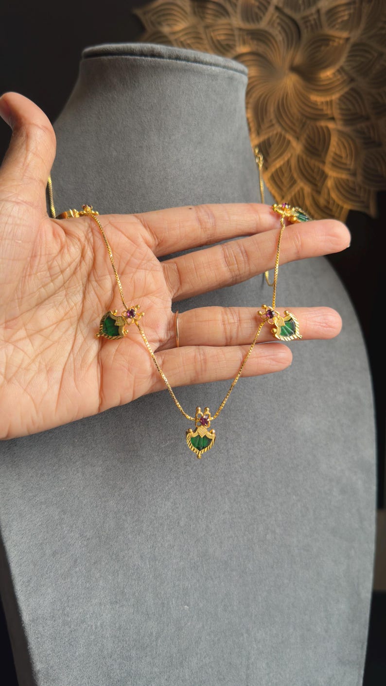 May include: Gold-coloured necklace with green and red gemstone accents. The necklace features a central pendant and two side pendants, all with a heart-shaped green stone and a small red stone. The necklace is held against a grey background.
