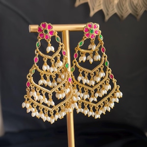 May include: A pair of gold-toned chandelier earrings featuring a tiered, chevron-shaped design. Each earring is adorned with small, vibrant pink and green enamel flower accents at the top and numerous dangling white seed pearls throughout.
