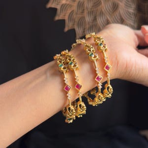 Può includere: Set di quattro braccialetti d'oro con disegni intricati, con pietre preziose rosse e verdi. I braccialetti sono adornati con pietre bianche e hanno un motivo a pavone.