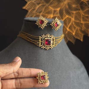 Könnte beinhalten: Ein goldfarbenes Schmuckset mit Halskette, Ohrringen und einem Ring. Jedes Schmuckstück hat einen quadratischen, rubinroten Mittelstein, der von kleineren, klaren Steinen umgeben ist. Die Halskette hat mehrere Stränge.
