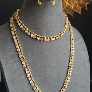 Puede incluir: Un conjunto de joyas de color dorado con un collar y pendientes a juego. El collar es un diseño de doble hebra con un patrón floral. Los pendientes son pequeños tachuelas con un diseño floral similar. Las joyas se exhiben en un maniquí gris.