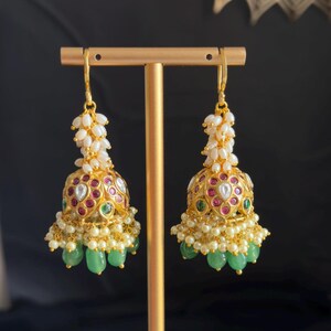 Puede incluir: Un par de pendientes jhumka dorados con un diseño en forma de campana adornado con pequeñas perlas blancas, piedras rojas y verdes incrustadas y cuentas verdes en forma de lágrima que cuelgan en la base, sobre un soporte de metal.