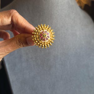 May include: Gold ring with a floral design. The ring has a central cluster of red and green gemstones, surrounded by white pearls and a border of small gold beads. The ring is held against a grey background.