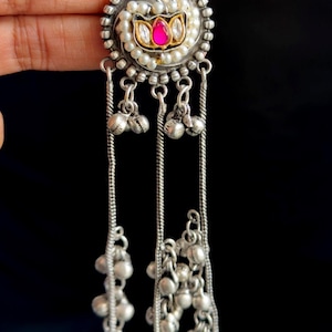 May include: Silver-toned dangle earrings with a circular design. The earrings feature a central pink gemstone surrounded by pearls and a decorative border. Dangling chains with small silver bells complete the design.