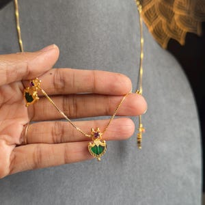 May include: A gold necklace and matching earrings set. The pendant and earrings feature a green gemstone with a gold frame and a small red gemstone accent. The jewellery is displayed on a grey surface.