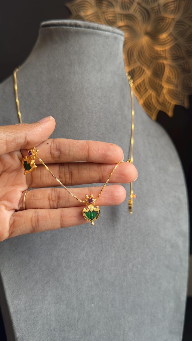 May include: A gold necklace and matching earrings set. The pendant and earrings feature a green gemstone with a gold frame and a small red gemstone accent. The jewellery is displayed on a grey surface.