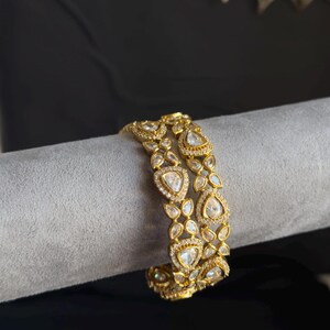May include: A pair of gold-toned bangles featuring a repeating pattern of teardrop-shaped clear stones surrounded by smaller pavé-set crystals. The metalwork has a textured finish, and the design consists of interconnected floral-like motifs.