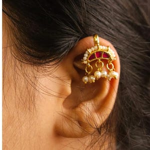 Könnte beinhalten: Ein goldfarbener Ohrschmuck mit einem roten und weißen Design, verziert mit kleinen Perlen und hängenden Perlenakzenten. Der Schmuck wird am Ohr getragen und zeigt seine komplizierten Details und seinen eleganten Stil.