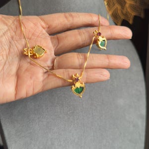 May include: A gold necklace and earrings set with heart-shaped pendants, adorned with green and red gemstones. The jewellery is displayed on a hand, highlighting the detailed design. The background is a neutral grey.