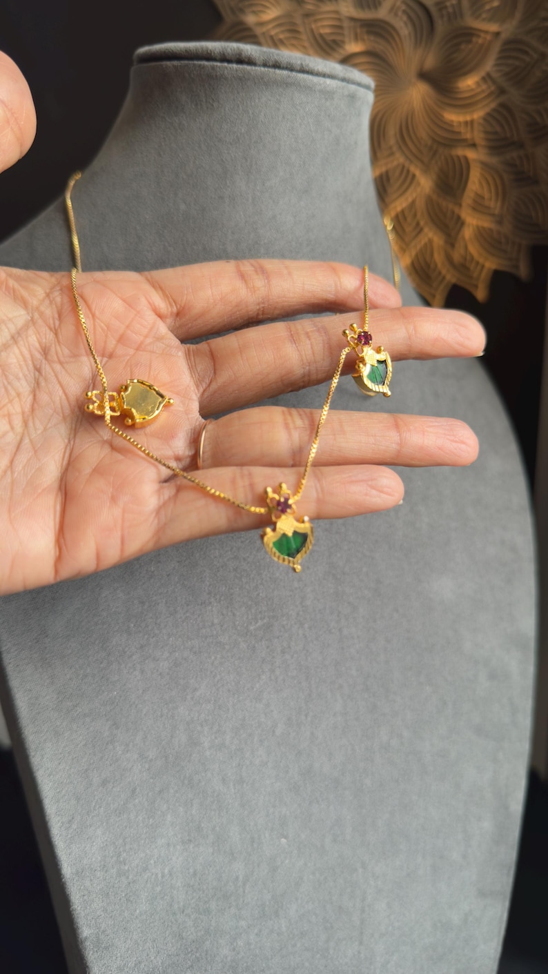 May include: A gold necklace and earrings set with heart-shaped pendants, adorned with green and red gemstones. The jewellery is displayed on a hand, highlighting the detailed design. The background is a neutral grey.