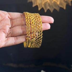 以下が含まれることがあります： 波状のテクスチャデザインの金色のバングルセット。 バングルは暗い背景に対して手に持たれており、右上の隅にはぼやけた装飾パターンがあります。 バングルは金属製のように見えます。