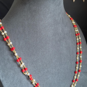 May include: A double-strand necklace with red and white beads, strung on a gold-colored chain. The necklace is displayed on a gray mannequin bust. The beads are small and evenly spaced, creating a delicate and elegant look.