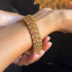 May include: Gold bracelet with multiple rows of gemstones. The bracelet features clear, red, and green stones, creating a vibrant design. The jewellery is worn on a wrist, showcasing its intricate details and craftsmanship. A piece of elegant jewellery.