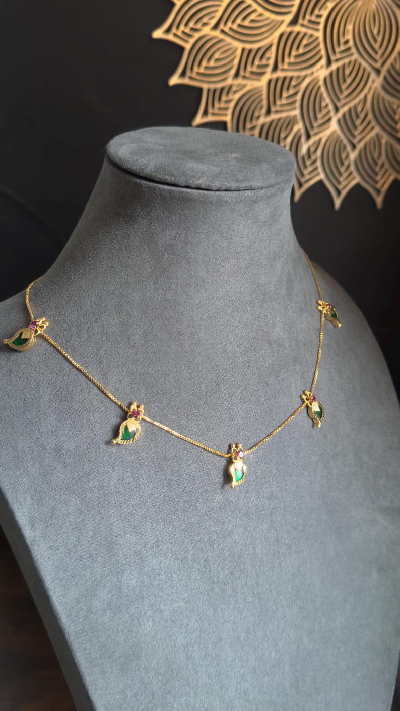 May include: A gold necklace with leaf-shaped pendants. Each pendant has a green centre stone and a small red stone. The necklace is displayed on a grey mannequin.