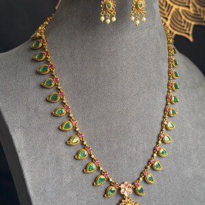 May include: A gold-toned necklace and earring set. The necklace features green teardrop-shaped stones and small red stones, with a pendant of a larger green stone and pearls. The earrings have a similar design.