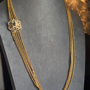 May include: A gold-toned necklace with multiple strands. The necklace features a floral pendant with a central green stone and surrounding clear stones. The necklace is displayed on a gray mannequin.