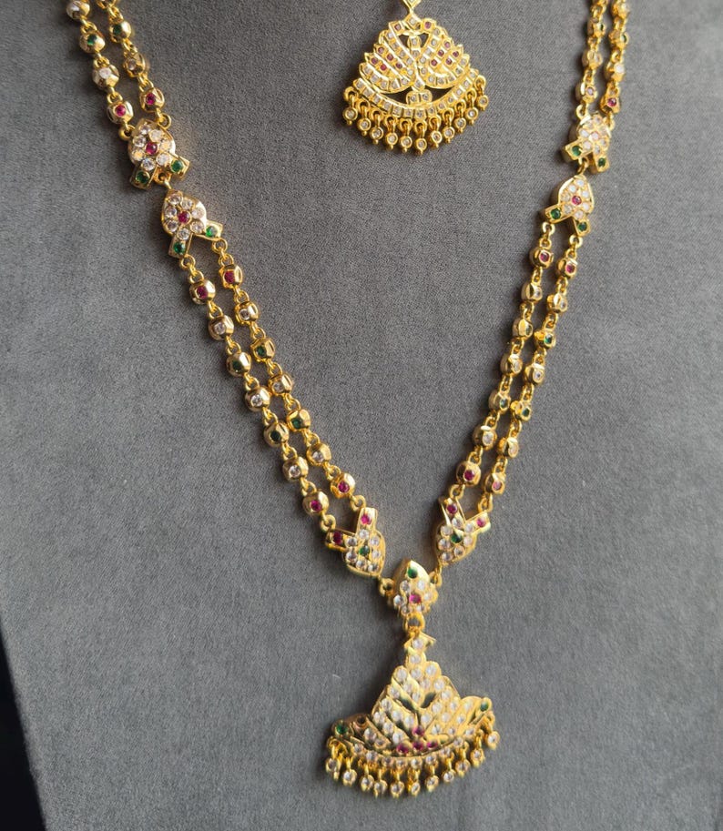 May include: A gold-toned necklace featuring a double-strand chain with repeating floral-shaped links set with small white, red, and green stones. The necklace includes a large, fan-shaped pendant and a matching smaller pendant piece.