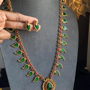 May include: A gold-toned necklace and matching earrings set. The necklace features green leaf-shaped pendants with red accents, and a large central pendant. The earrings mirror the leaf design. The jewelry is displayed on a gray surface.