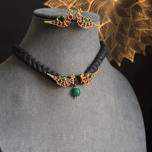 Puede incluir: Un collar de oro con un cordón trenzado negro y un colgante con dos pájaros con piedras preciosas rojas y verdes. El colgante tiene una sola piedra preciosa verde que cuelga de la parte inferior.