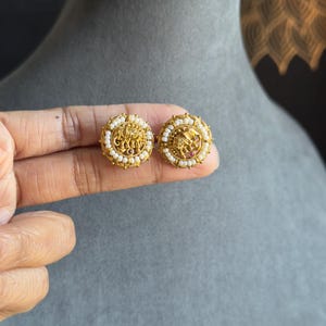 May include: A pair of gold-colored stud earrings. Each earring features a central design with an elephant motif, surrounded by small white beads. The earrings are held up by a hand against a gray background.