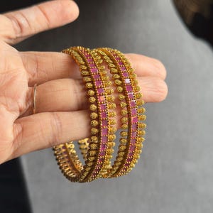 Puede incluir: Brazaletes dorados con un patrón repetitivo de pequeñas cuentas redondas y piedras preciosas rosas cuadradas. Las joyas se sostienen en una mano, mostrando el intrincado diseño y el contraste de color. Los brazaletes son de estilo tradicional.