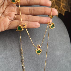 May include: Gold-coloured necklace with decorative pendants featuring green and red details. The necklace is displayed against a grey backdrop, highlighting its intricate design. The chain has a delicate, linked design, showcasing the craftsmanship.