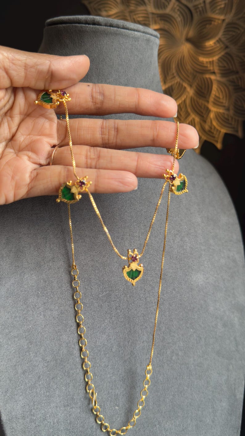 May include: Gold-coloured necklace with decorative pendants featuring green and red details. The necklace is displayed against a grey backdrop, highlighting its intricate design. The chain has a delicate, linked design, showcasing the craftsmanship.