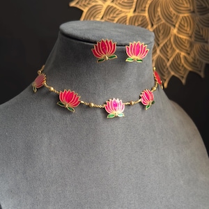 Puede incluir: Un conjunto de collar y pendientes dorados. La joyería presenta colgantes de flores de loto rosas con detalles verdes. El collar y los pendientes se exhiben sobre un maniquí gris sobre un fondo oscuro.