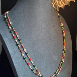 May include: A gold-toned necklace with two strands of small, colorful beads. The beads are red, green, and white, interspersed along the gold chain. The necklace is displayed on a gray mannequin.