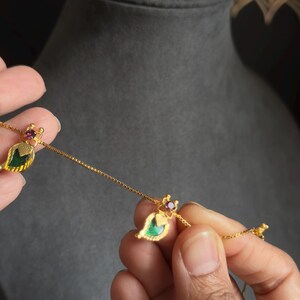 May include: A gold necklace with leaf-shaped pendants. Each pendant has a green centre stone, a small red gem, and gold detailing. The necklace is held against a dark grey background.