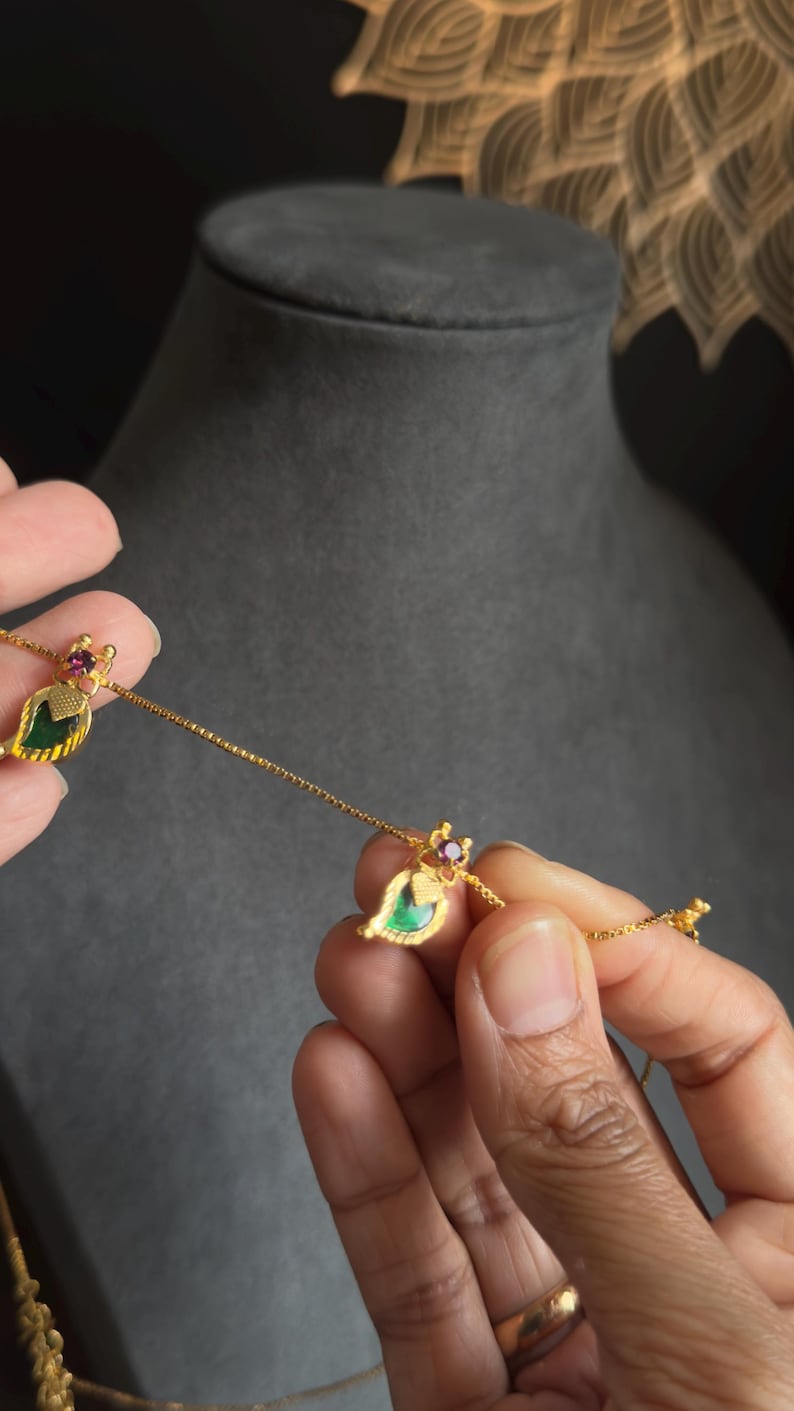 May include: A gold necklace with leaf-shaped pendants. Each pendant has a green centre stone, a small red gem, and gold detailing. The necklace is held against a dark grey background.