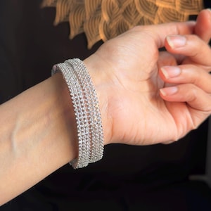 Puede incluir: Una pulsera plateada con múltiples filas de pequeñas piedras brillantes. La pulsera se lleva en la muñeca, con un fondo oscuro y un objeto decorativo de color dorado en el fondo.