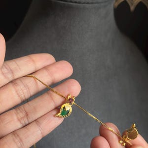May include: A gold necklace with a leaf-shaped pendant. The pendant has a green gemstone and a small red gemstone. The necklace is held by a person's hand, against a dark grey background.