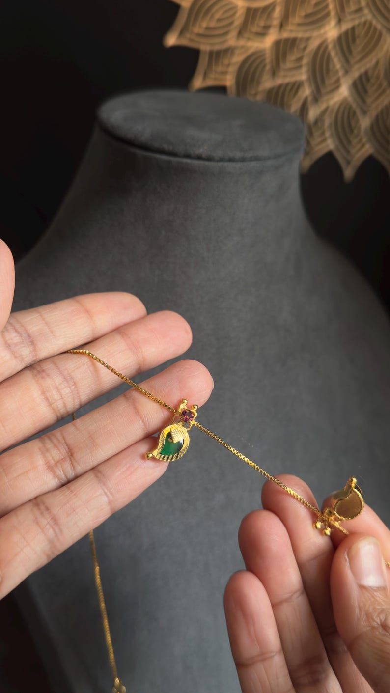 May include: A gold necklace with a leaf-shaped pendant. The pendant has a green gemstone and a small red gemstone. The necklace is held by a person's hand, against a dark grey background.